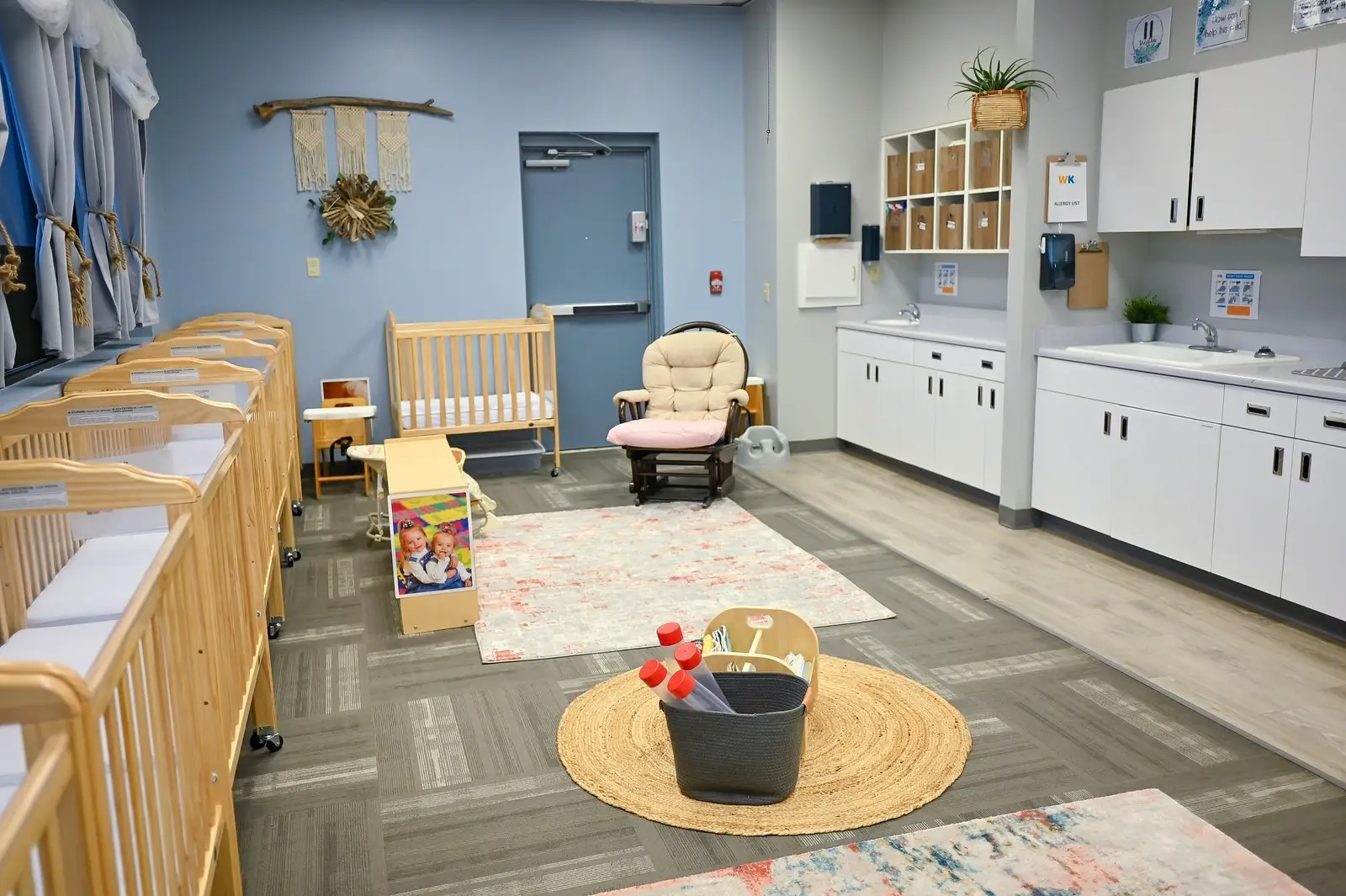 A brightly lit infant daycare room features wooden cribs lined against the left wall. A wooden rocking chair, two cozy rugs, and a bin of toys add charm. White cabinets with sinks grace the right side. Decorations include colorful wall hangings, a plant, and a child-sized kitchen set—a perfect choice among daycares nearby.