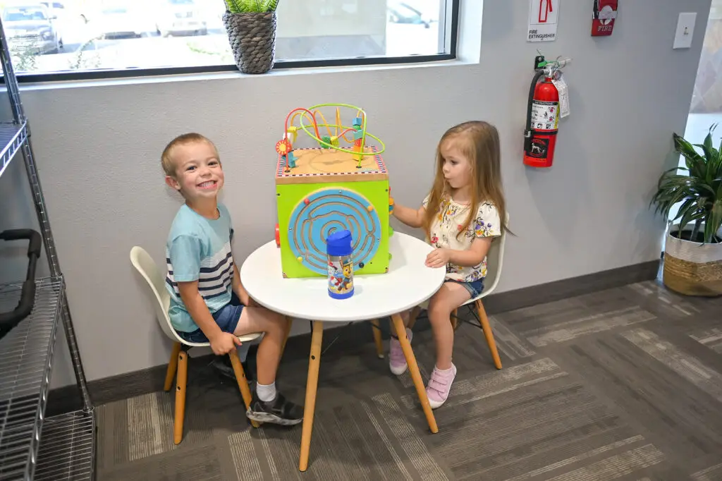 Two young children are seated at a small round table, playing with a colorful activity cube in a cozy room that resembles those found in childcare near me. The boy on the left smiles at the camera, while the girl focuses intently. The space includes a window, fire extinguisher, and potted plant against gray walls.