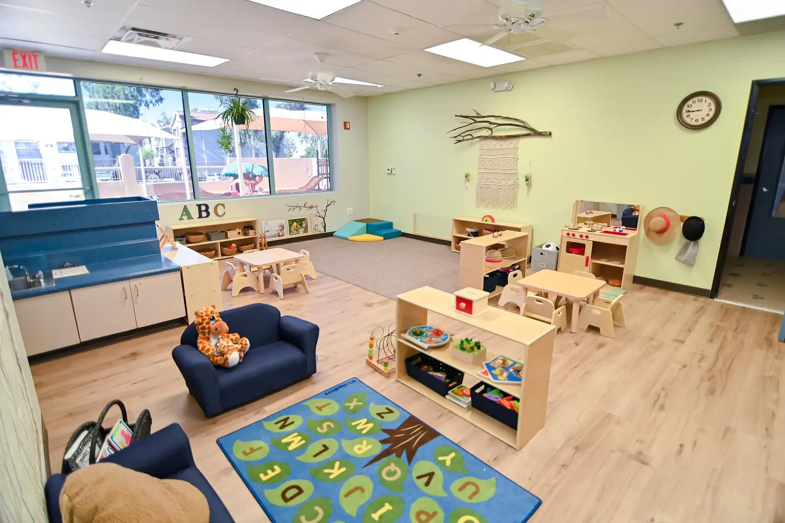A brightly lit preschool classroom with wooden floors and play stations exudes warmth. It features a blue armchair with a teddy bear, small tables and chairs, and an alphabet mat. Light green walls adorned with decor complement large windows for natural light—a charming scene at any pre-k near me.