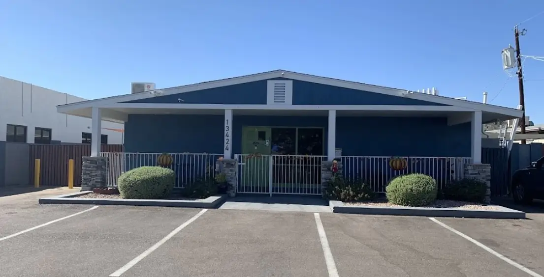 This charming single-story building with blue siding and white trim could be a perfect spot for families seeking childcare near me. The entrance features a welcoming porch with columns and stone accents, and the pathway, flanked by shrubs, leads to a fenced area. Parking spaces and a utility pole are visible in the foreground against a clear sky backdrop.