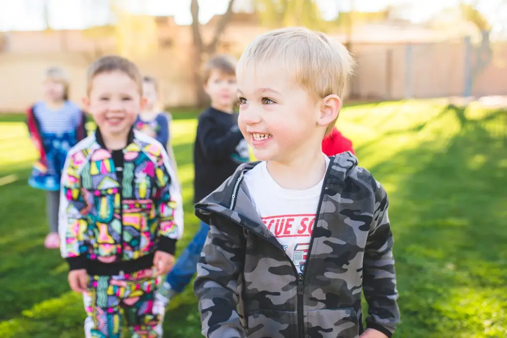 Children are playing outdoors on grass. The focus is on a boy in a camouflage jacket, smiling and looking to the side. Nearby, pre-k friends join the fun, with one child in a colorful outfit smiling directly at the camera. Sunlight casts soft shadows as they play near their daycare.