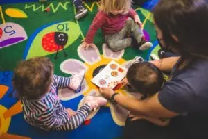Children gather with a caregiver on a vibrant ladybug-themed rug. The caregiver, positioned to the right, shares a book filled with food illustrations as the kids remain focused and engaged. One child sports striped clothing, another wears a red shirt—a perfect snapshot of pre-k learning near me.