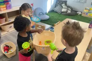 Three children are gathered around a basket of colorful toys, including rattles and tambourines, in a brightly lit preschool classroom. A wooden table is nearby, and shelves with books and cushions are in the background. The floor is partially carpeted with green turf, reflecting the warmth of daycares nearby.