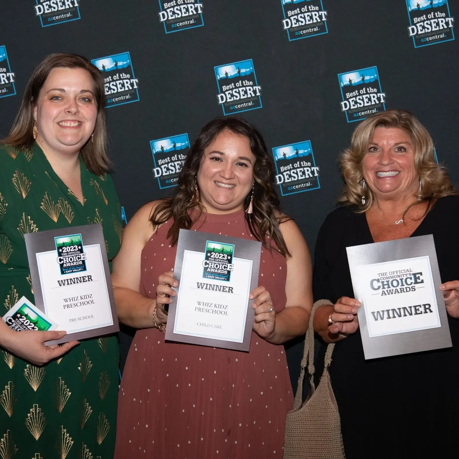 Three women smile and hold award plaques at a Best of the Desert event. Dressed in semi-formal attire, they stand before a step-and-repeat banner. Two plaques highlight Winner 2023 Choice Awards, celebrating excellence in childcare and preschools near me categories.