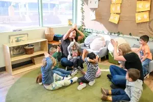 A diverse group of young children and two adults sit in a circle on a green rug at a local childcare near me. They are engaged in a playful activity, raising their hands above their heads. The room is adorned with drawings and plants, as sunlight streams through the window in the background.