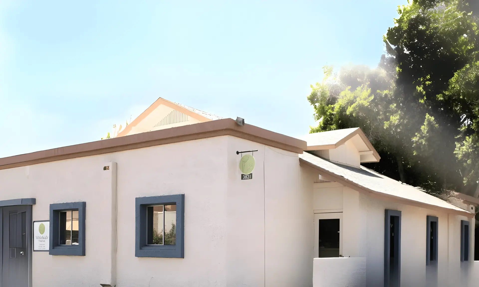 A small, white stucco building with blue-trimmed windows and a wooden roof is shown, resembling charming childcare near me. A circular sign with a leaf design hangs by the entrance. Lush green trees are visible in the background under a clear blue sky. A plaque is mounted on the wall near the door.