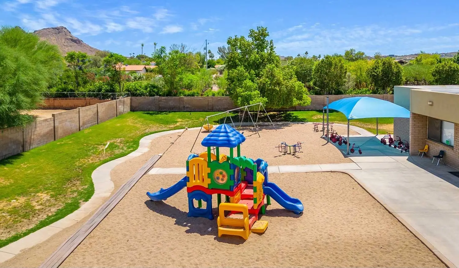 Colorful playground equipment with slides and climbing sections on sandy ground, ideal for preschools near me. Nearby, a swing set and shaded area with tables provide a perfect spot for daycares nearby. Surrounding greenery and a mountain in the background enhance the outdoor setting under a clear blue sky.